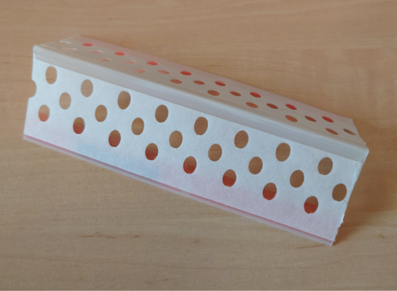 Vit perforerad plastprofil med röd tejp, liggande på ett träbord.